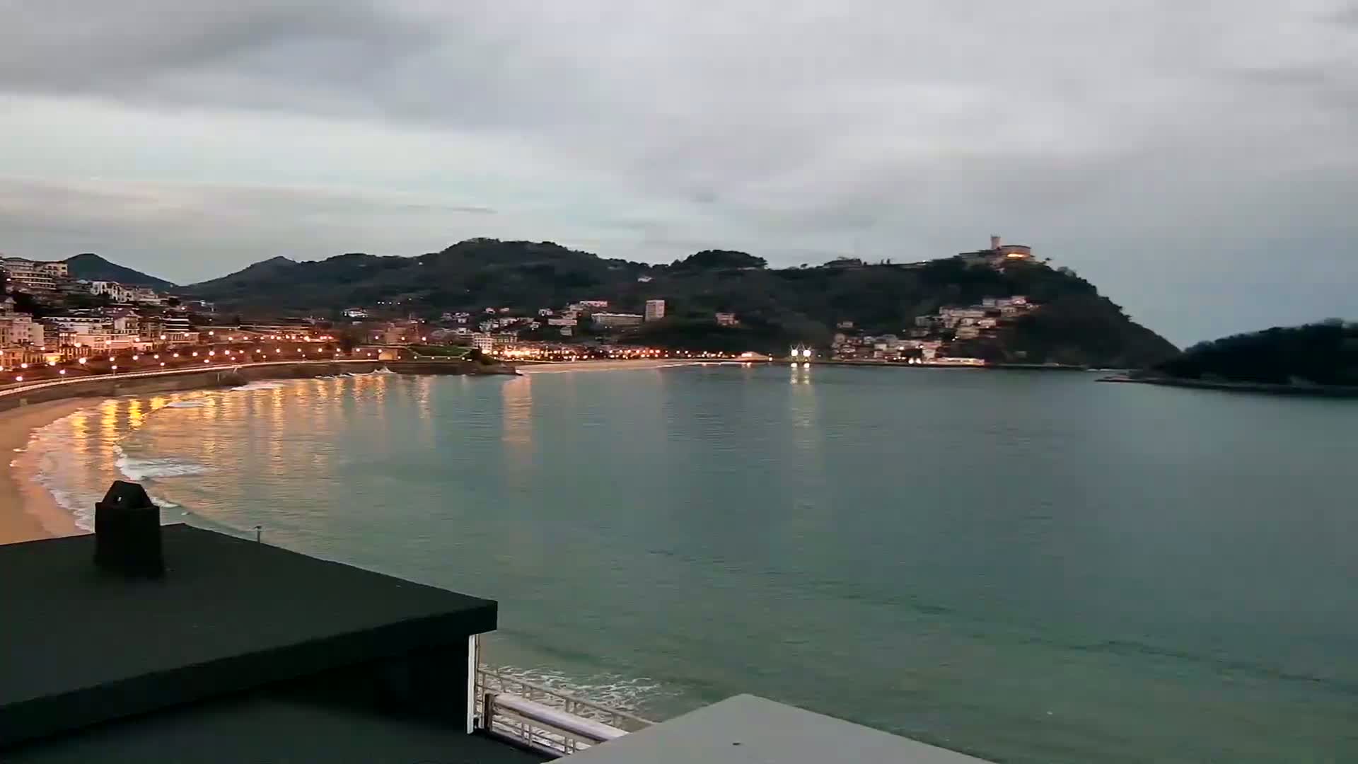 A nighttime view of a coastal town nestled between hills, with a calm bay reflecting the lights of the buildings under an overcast sky.