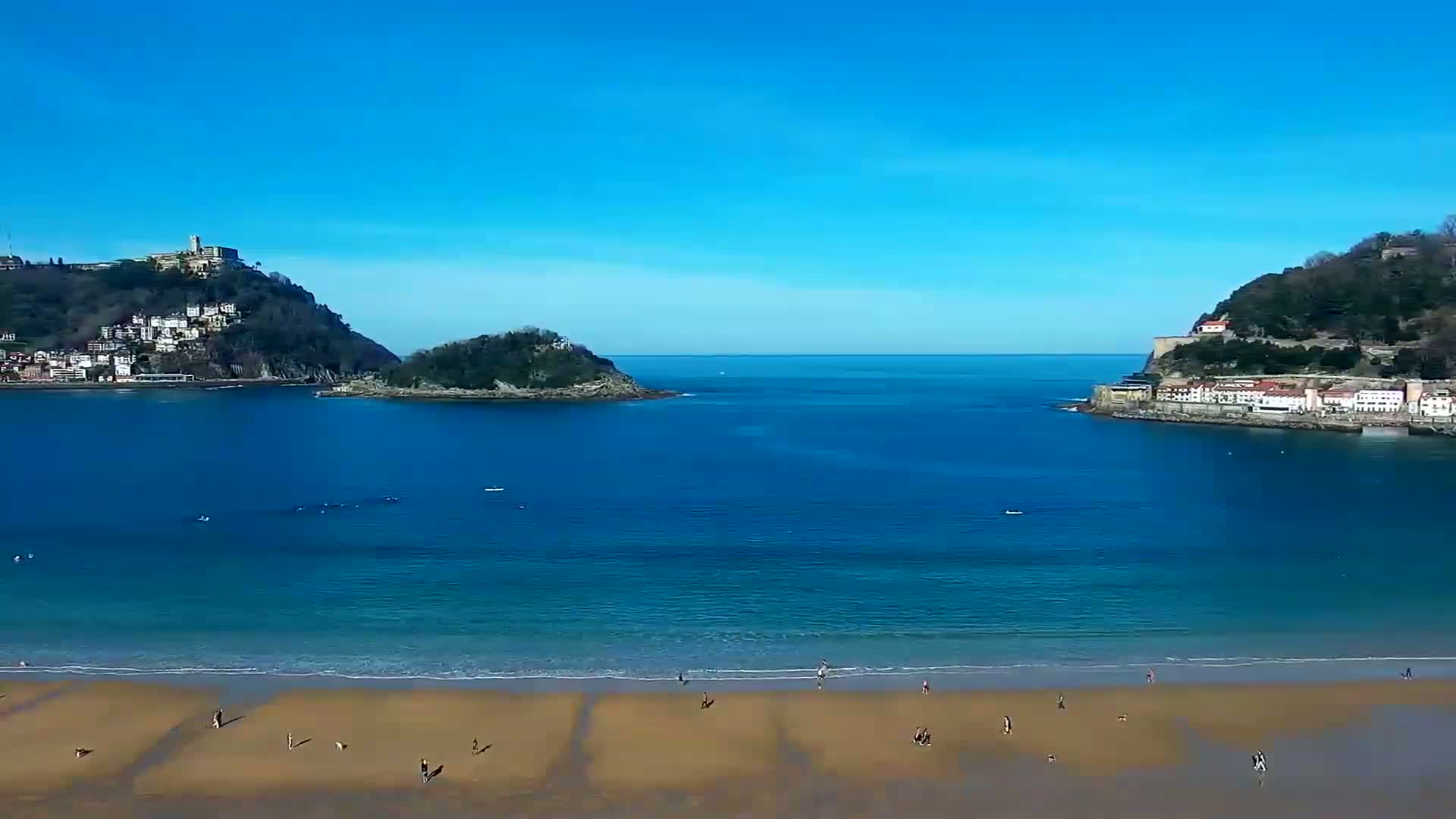 A sandy beach with scattered people stretches along a calm, blue ocean, bordered by hills topped with buildings under a clear blue sky.