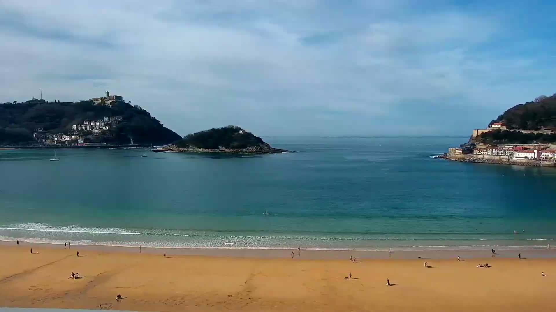 A sandy beach with scattered people is nestled between two hillsides, one featuring buildings, under a partly cloudy sky, with calm, clear blue water separating the landmasses.