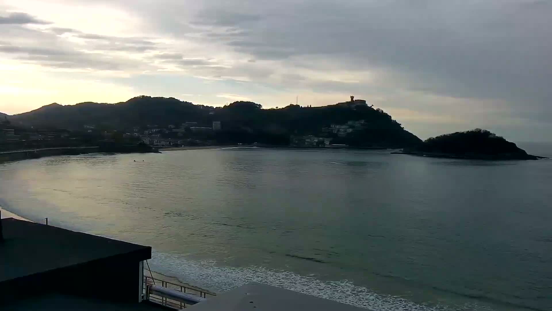 A calm bay with gentle waves laps a sandy shore, backed by hills and a small island under a cloudy, dusky sky.