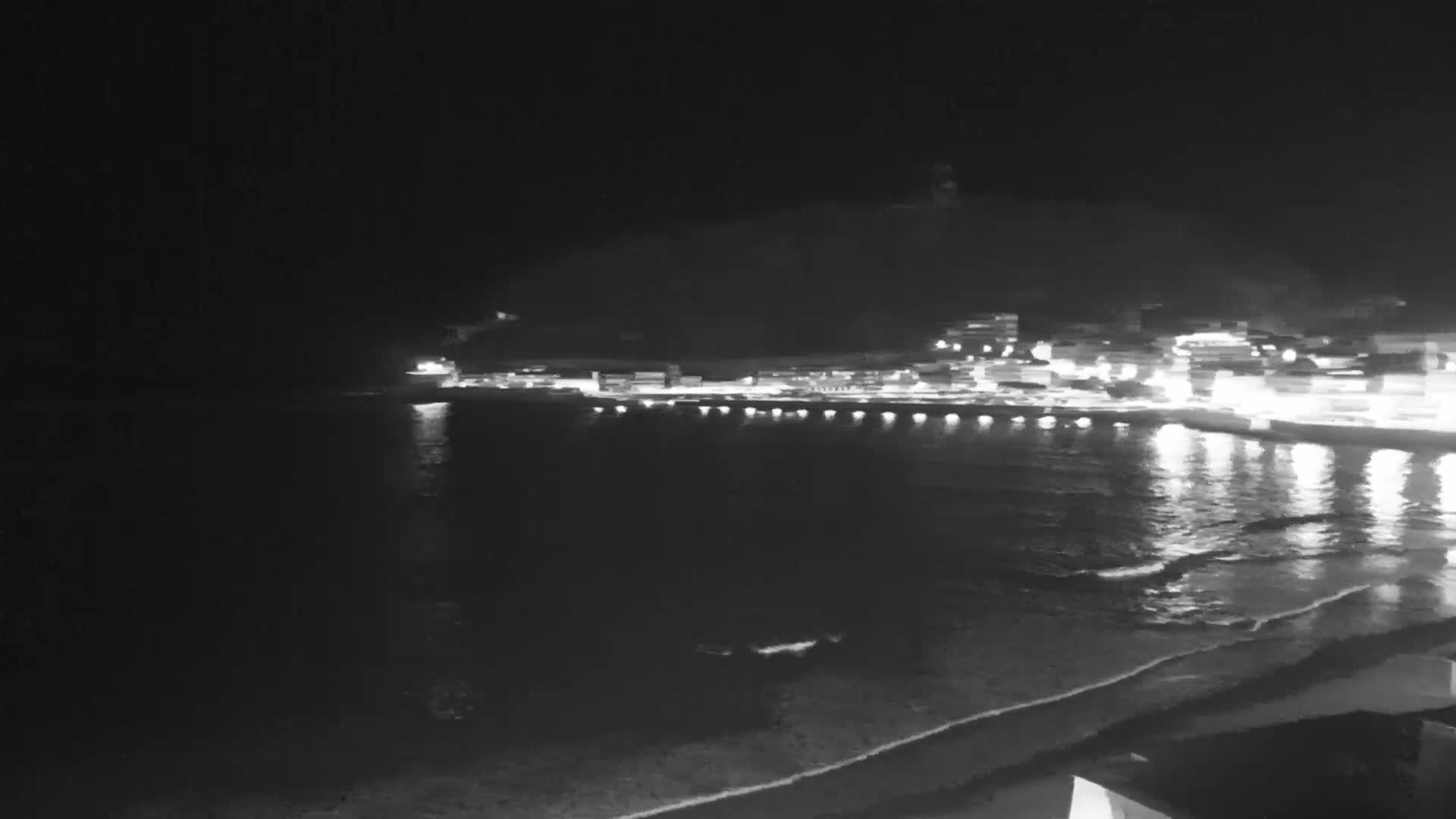 A nighttime view of a dark, calm ocean reflecting the lights of a coastal town situated at the base of a hill.