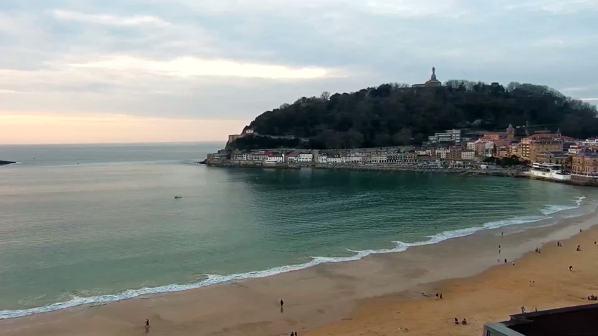 A sandy beach with gentle waves laps the shore, facing a town nestled at the base of a tree-covered hill topped with a statue, all under a partly cloudy sky.