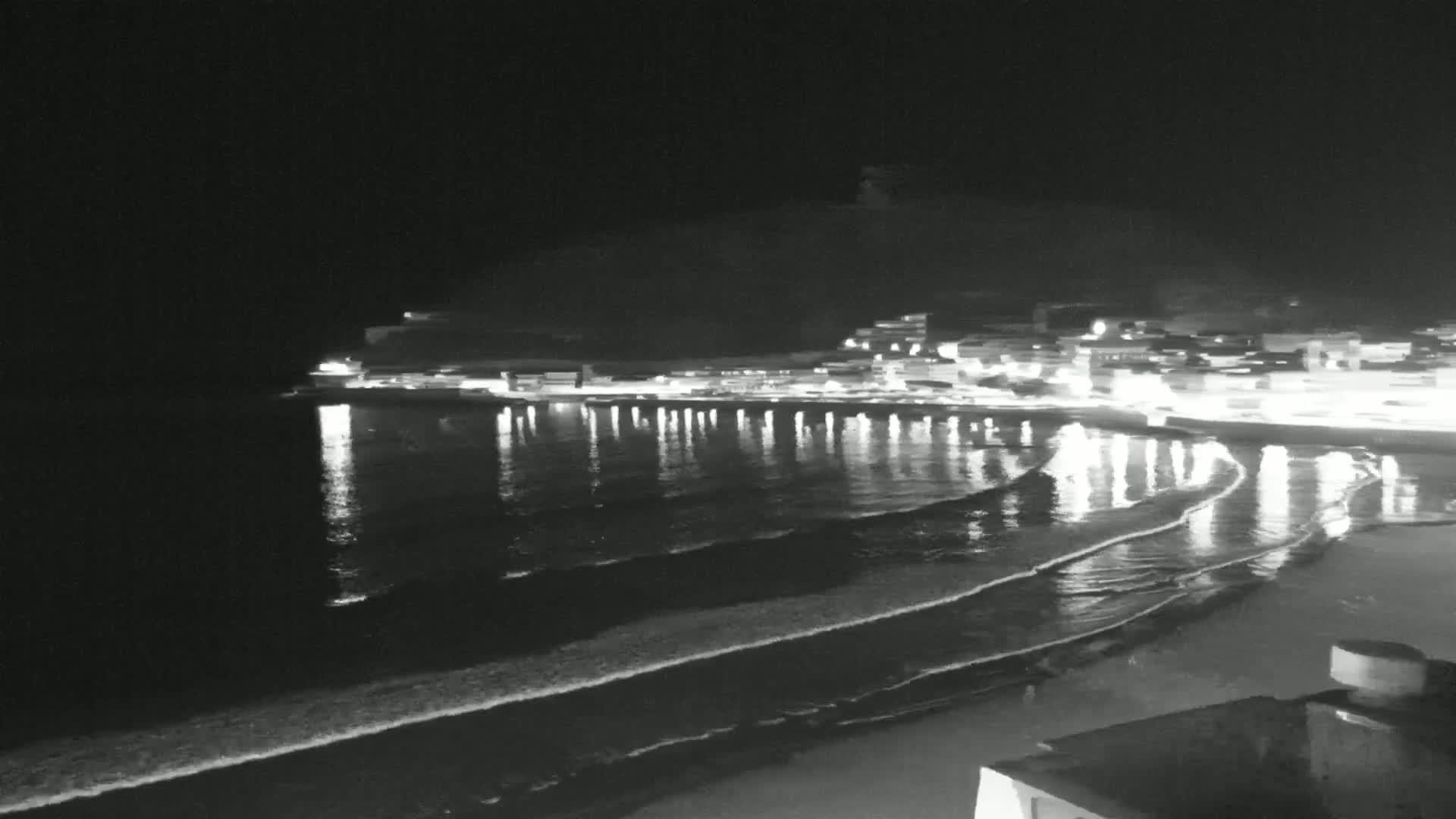 A nighttime view of a coastal town with lights reflecting on the gently lapping waves of a dark ocean.