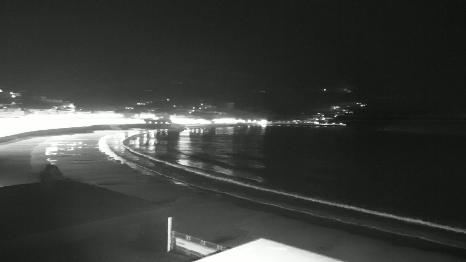 A nighttime view of a beach with gently lapping waves, illuminated by lights from buildings across the water.