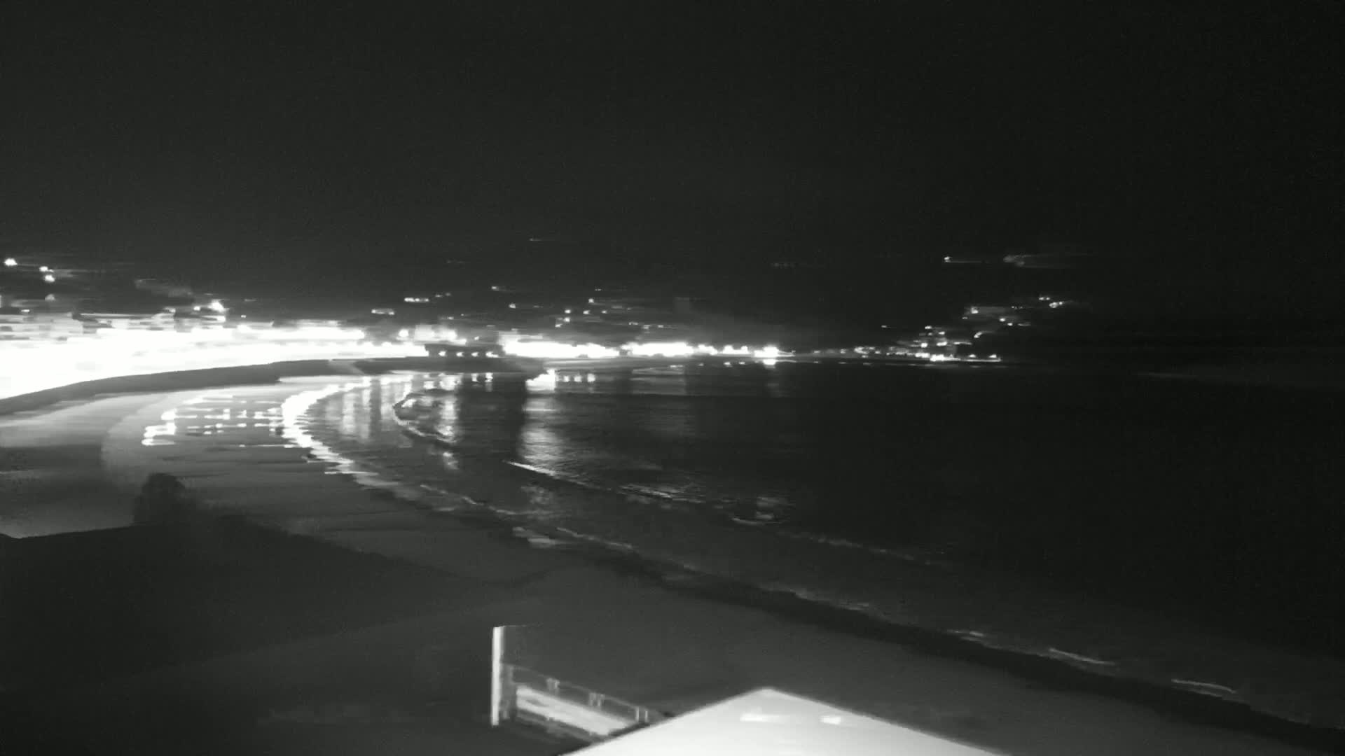 A dark, nighttime view of a beach with gently lapping waves and a town illuminated by lights in the distance.