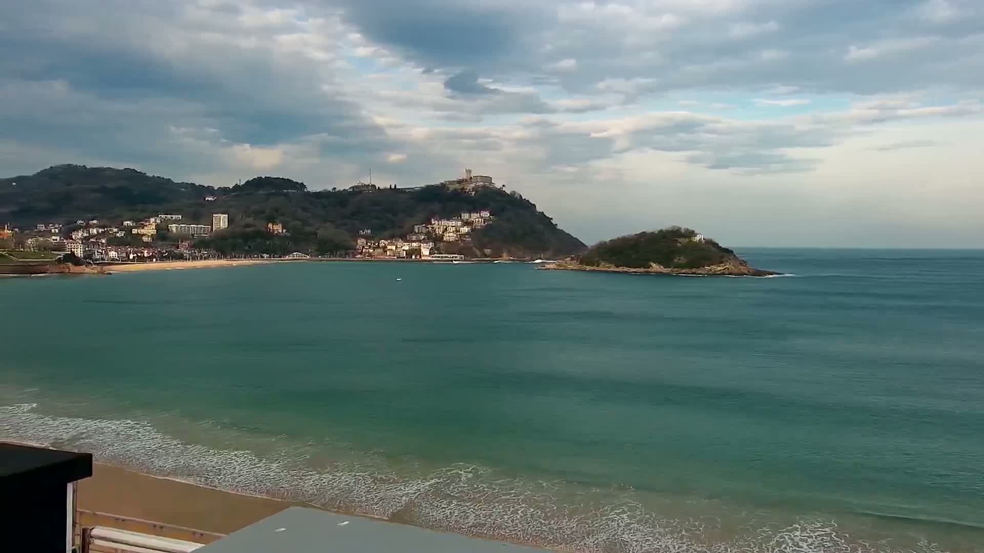 A sandy beach with gentle waves laps the shore, in front of a town nestled at the base of a hill, all under a partly cloudy sky.
