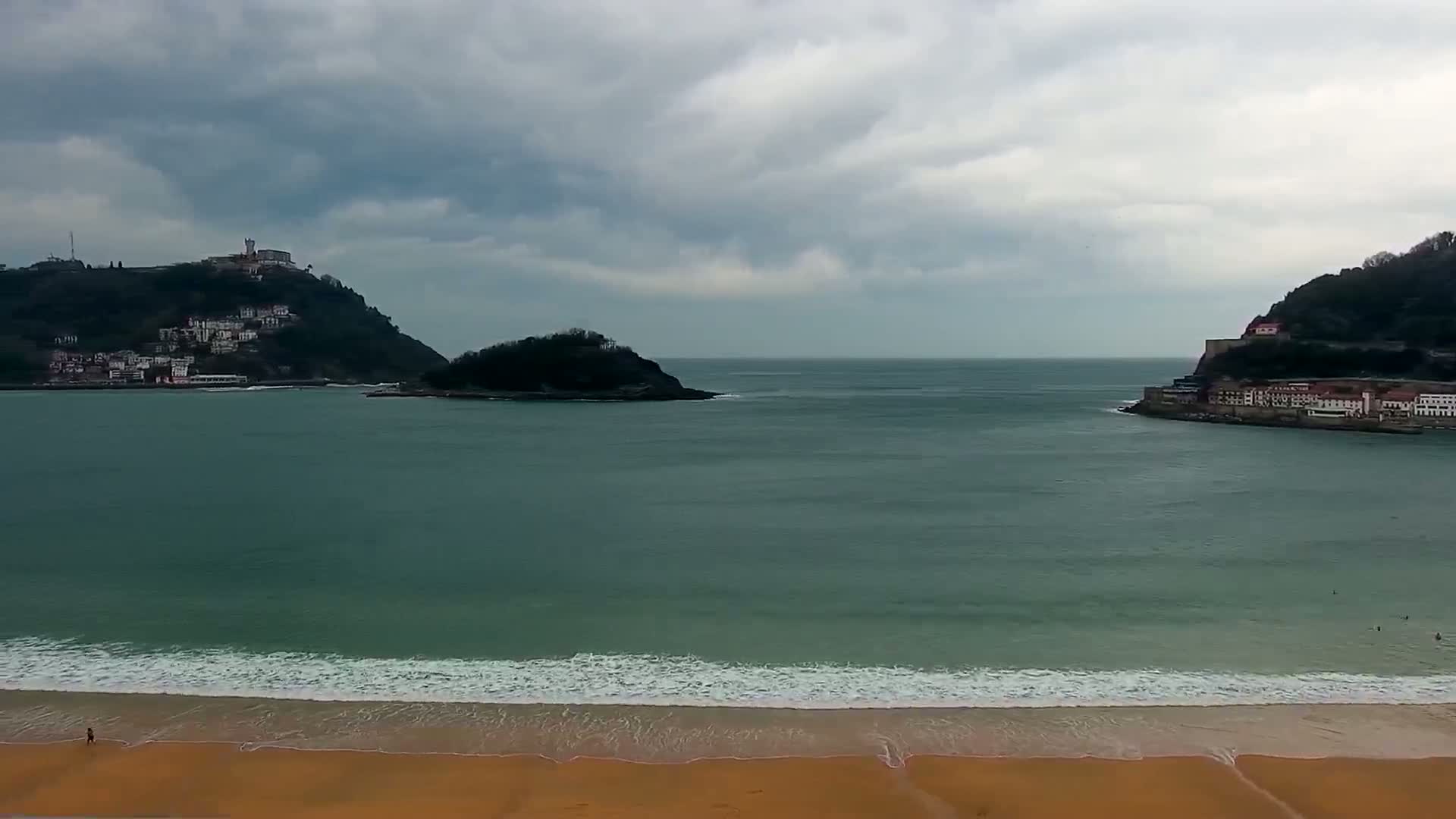 A sandy beach with small waves laps against the shore, overlooking a bay with two hills, one featuring buildings, under a cloudy sky.