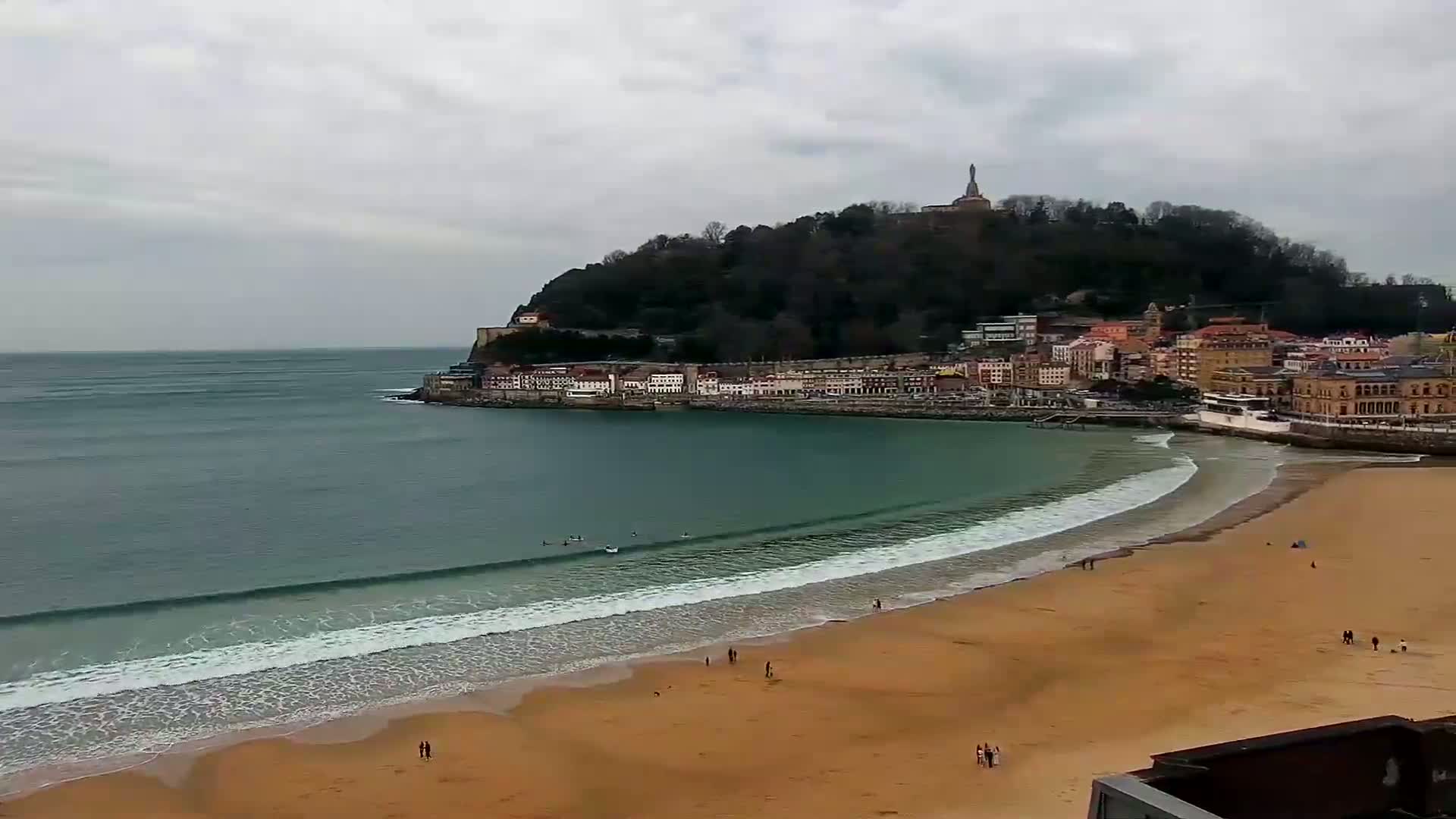 A sandy beach with gentle waves laps the shore, in front of a town nestled at the base of a tree-covered hill under an overcast sky.