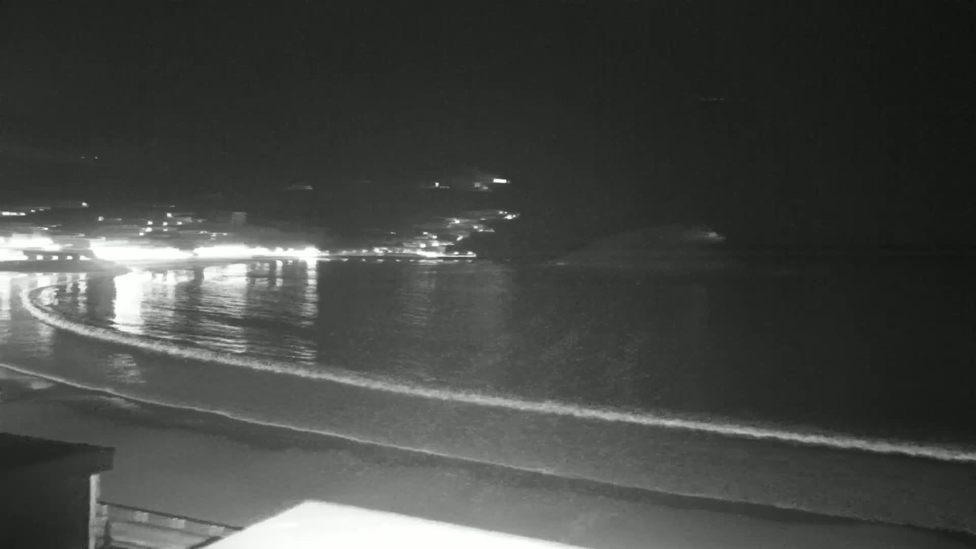 A nighttime view of a calm ocean with gentle waves lapping a beach, lit by the distant lights of buildings on a shore.