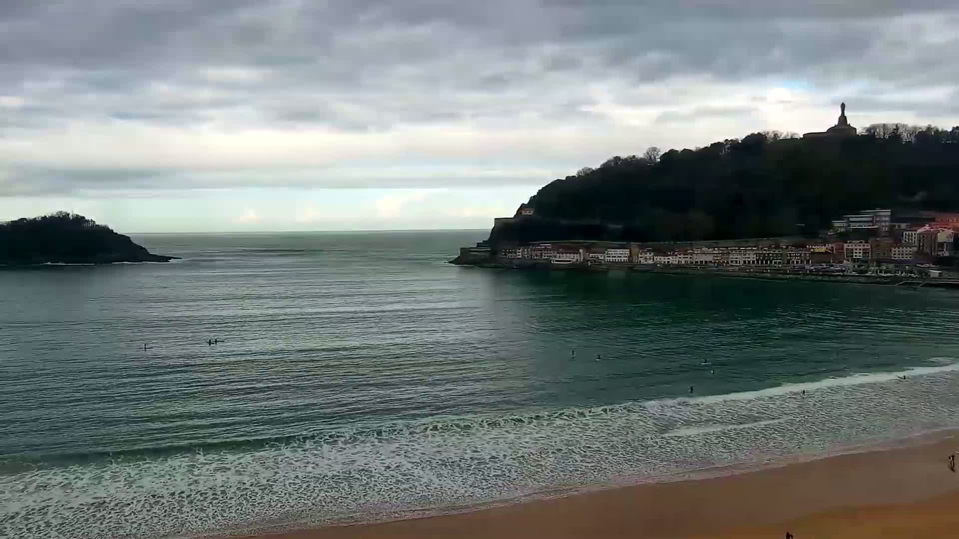 A sandy beach with small waves meets a calm bay, bordered by a town nestled against a hill topped with a statue, all under a cloudy sky.