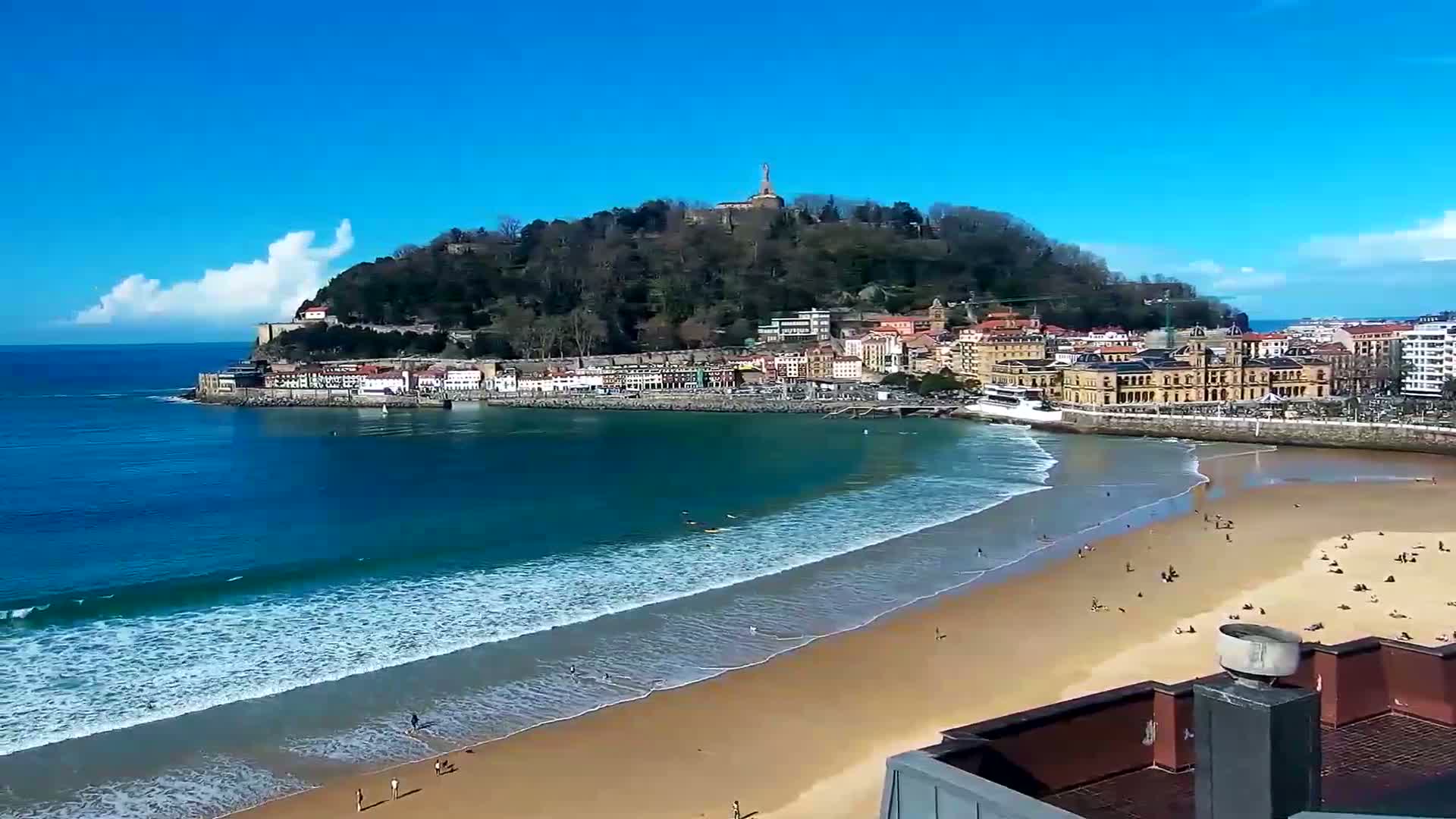 A sandy beach with gentle waves laps the shore beneath a town nestled against a tree-covered hill under a clear blue sky with a few fluffy clouds.