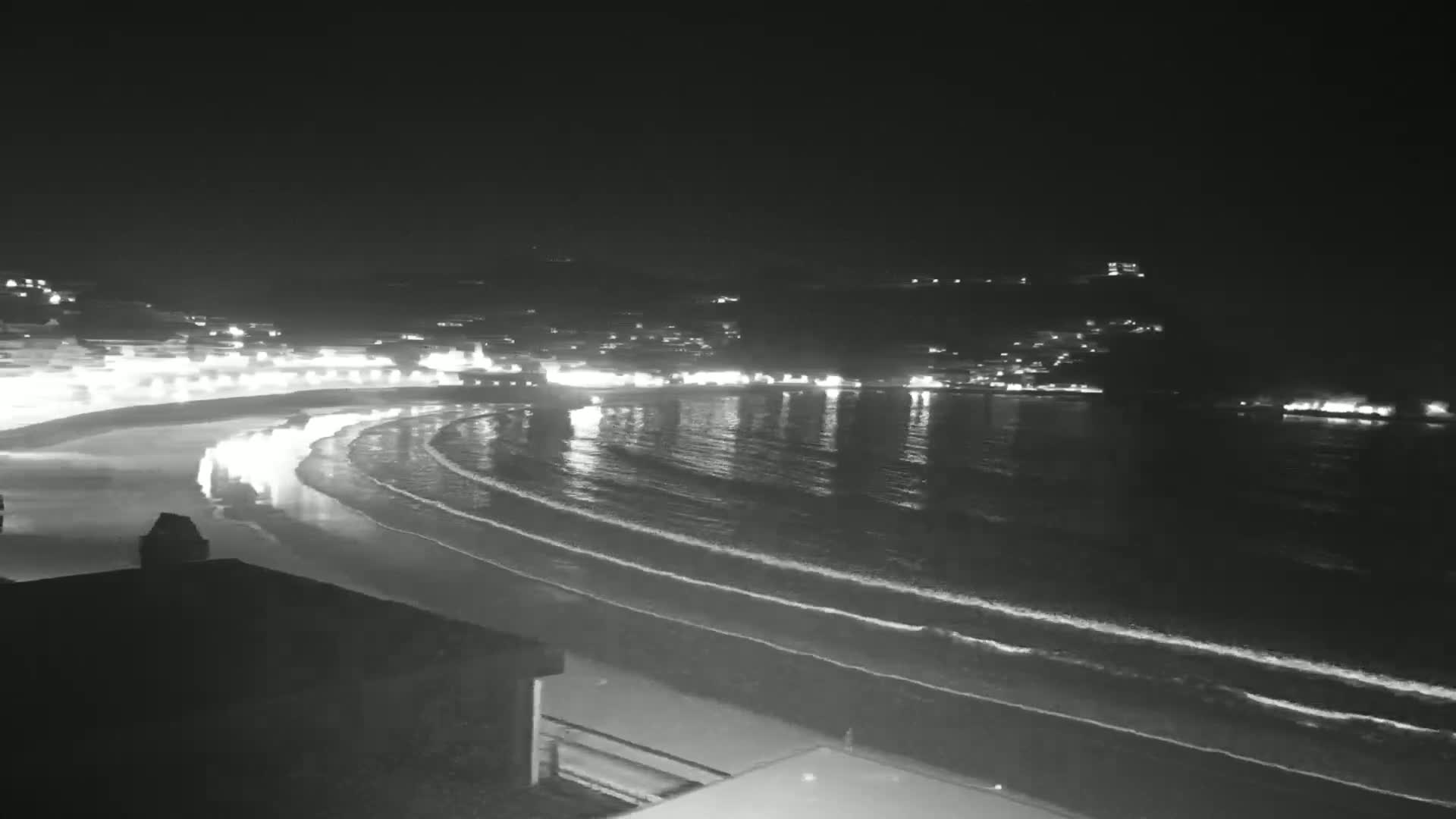 A nighttime view of a gently-sloping beach and calm ocean with a town's lights illuminating the shoreline in the background.