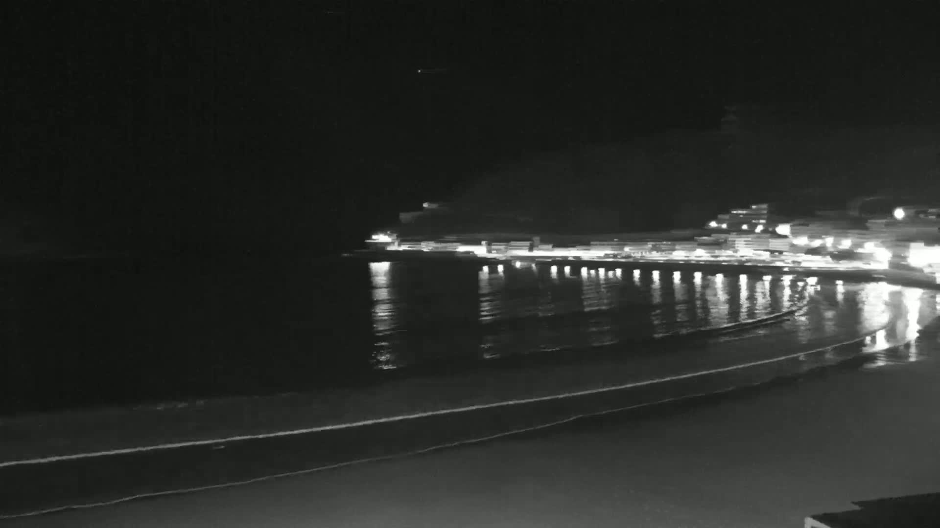 A nighttime view of a calm ocean bay with a town's lights reflected in the water, gently lapping waves on a dark sandy beach.