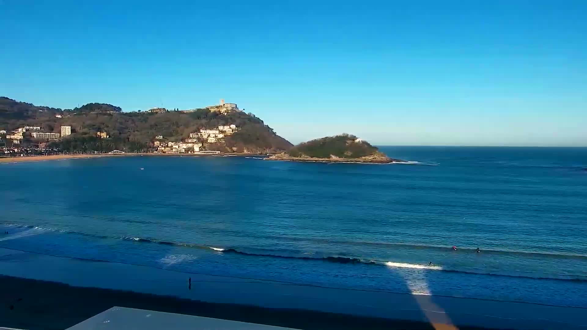 A sunny day shows a bay with gentle waves lapping a sandy beach, backed by a hill dotted with buildings and a smaller island offshore.
