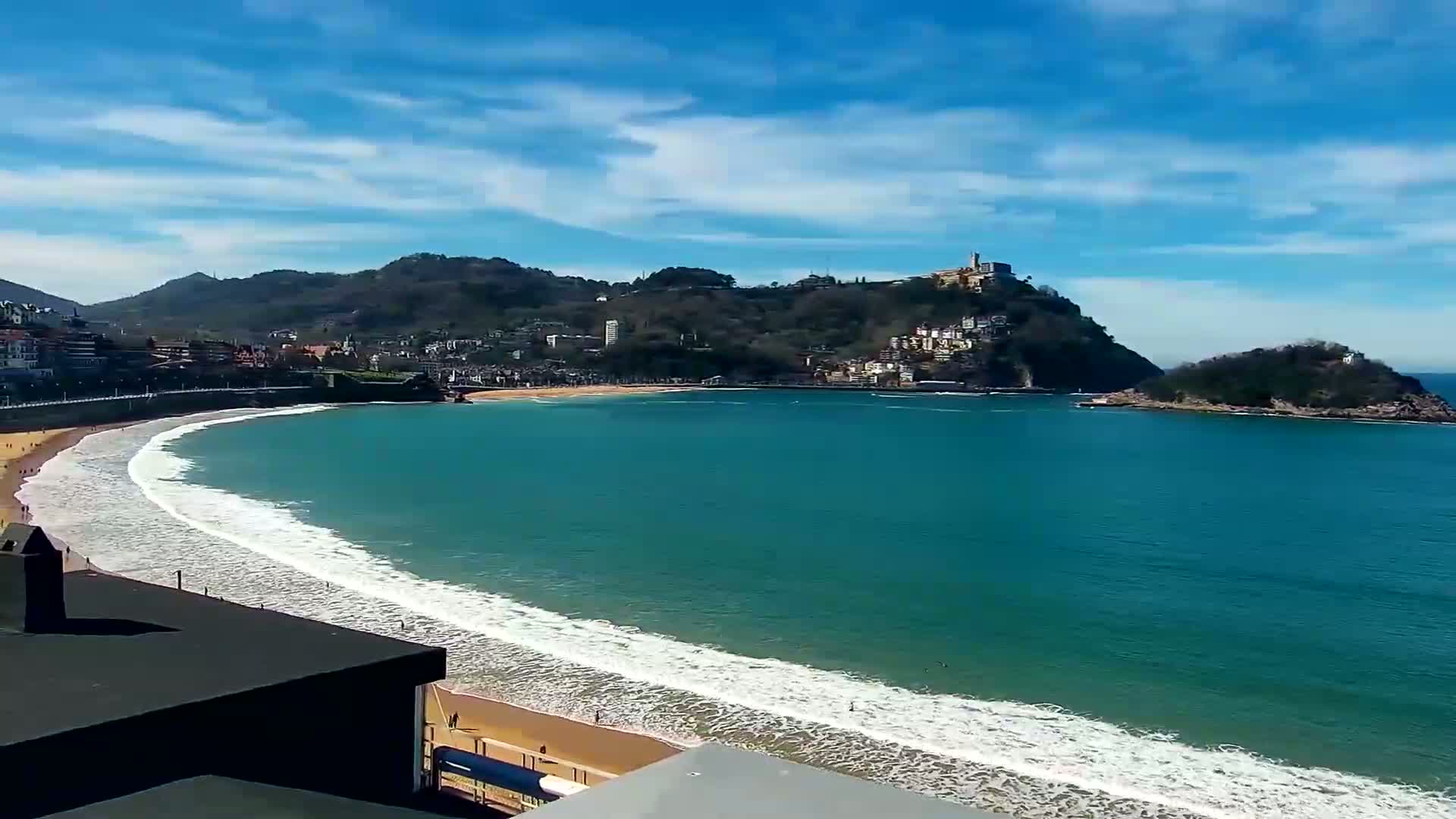 A sandy beach with gentle waves curves around a bay,  under a mostly sunny sky, with green hills and buildings rising in the background.