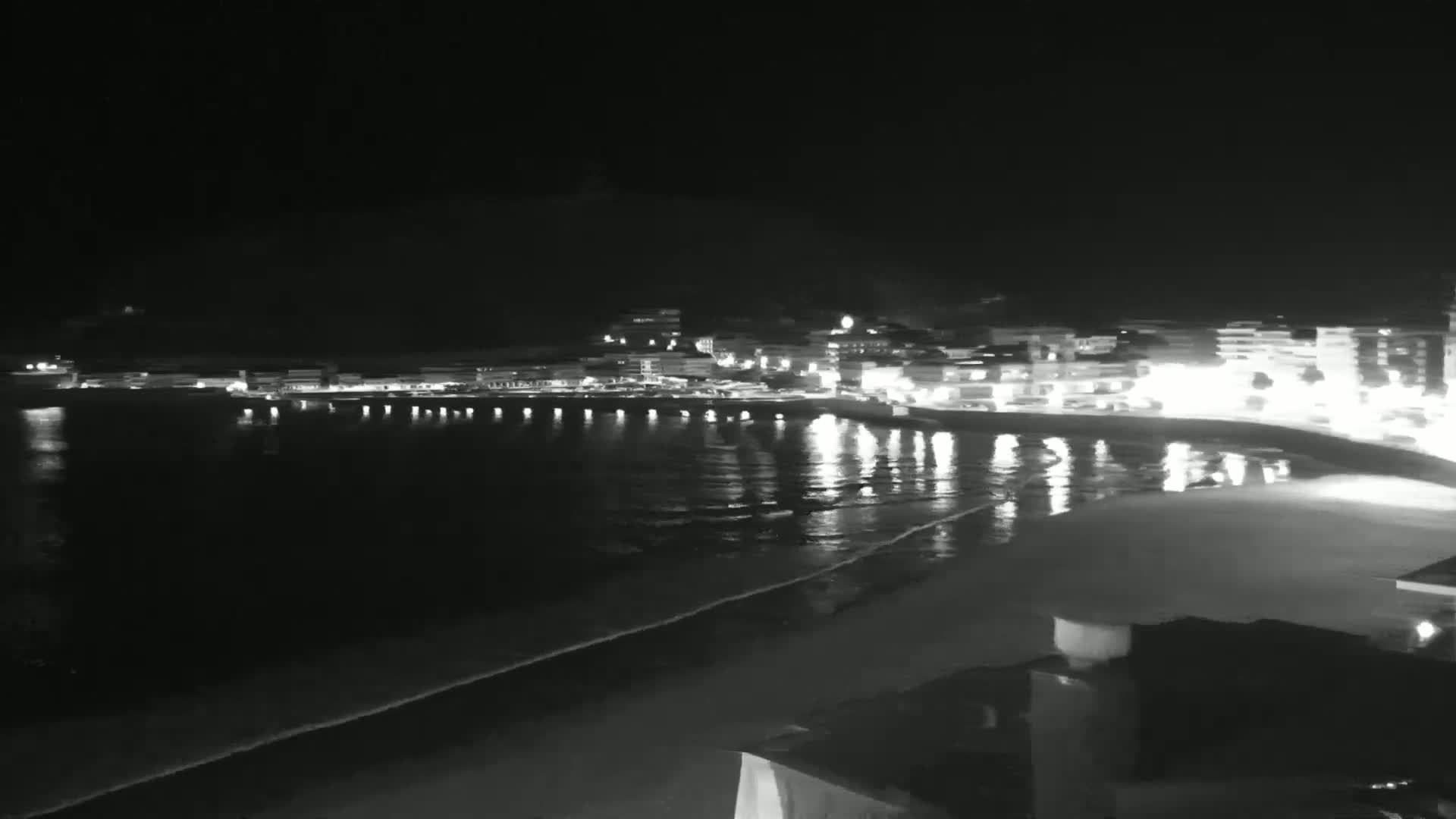 A nighttime view of a brightly lit coastal town with a calm, dark ocean reflecting the lights.