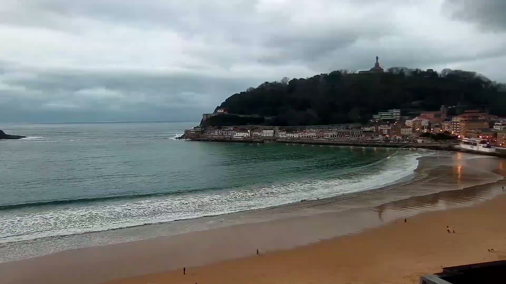 A sandy beach with gentle waves laps the shore under a cloudy sky, backed by a town nestled at the base of a hill topped with a statue.