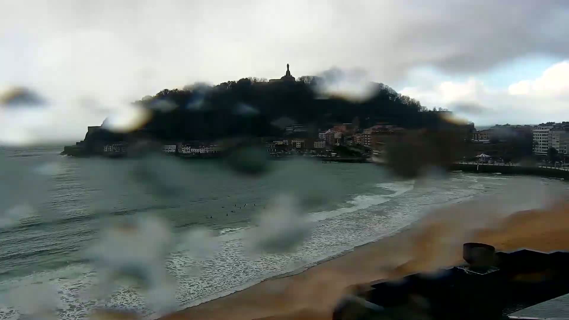 A rain-streaked view shows a beach with small waves lapping a sandy shore, buildings lining a coastline, and a hill with a statue on top in the distance.