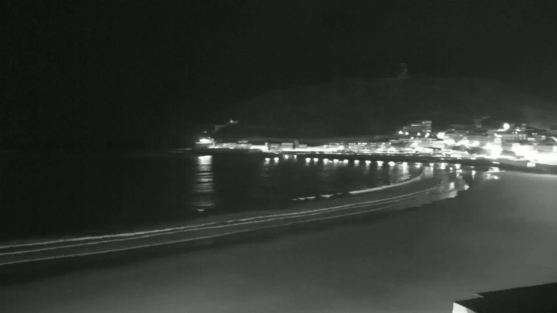 A dark, nighttime beach scene shows gently lapping waves on the shore, with the lights of a town reflecting on the water in the background.