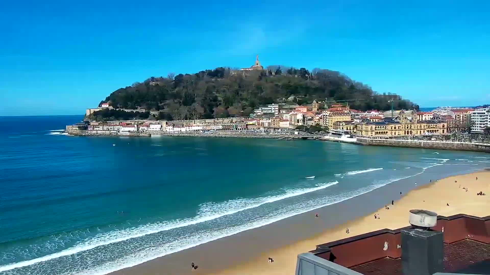 A sandy beach with gentle waves laps the shore in front of a town nestled at the base of a verdant hill under a clear blue sky.