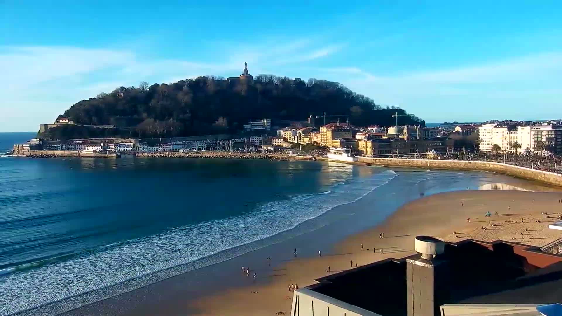 A sandy beach with gentle waves laps the shore under a clear blue sky, beside a town nestled at the base of a tree-covered hill topped with a monument.