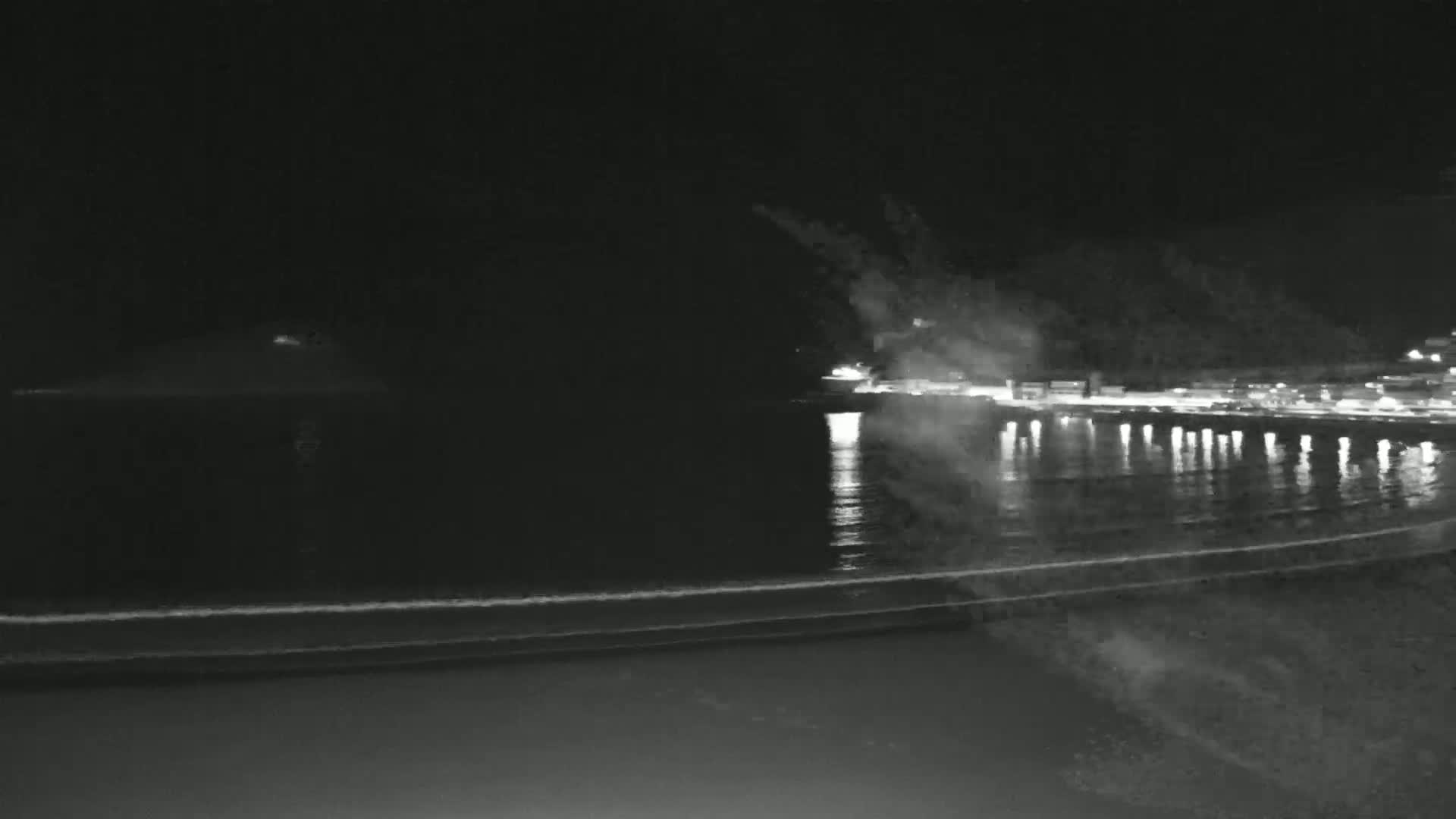 A nighttime view of a dark beach with gently lapping waves and a dimly lit pier across a body of water under a clear, dark sky.