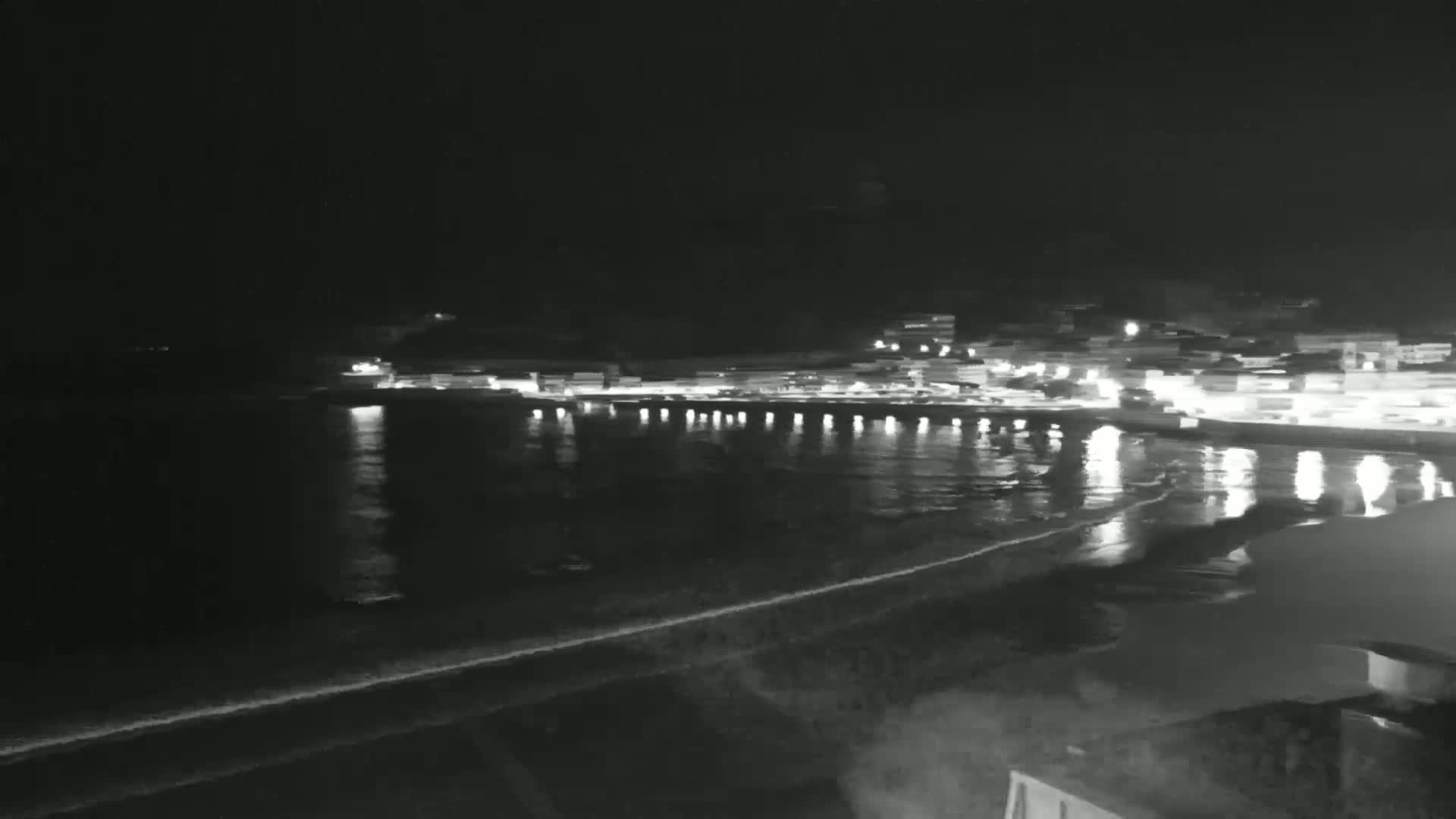 A nighttime view of a coastal town with lights reflecting on the calm water of a beach.