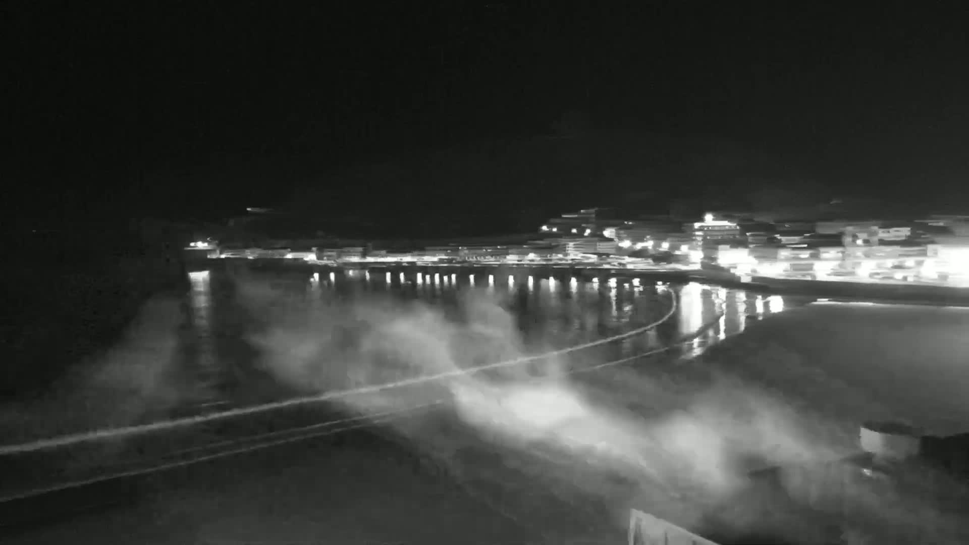 A nighttime, coastal town with lights reflecting on a turbulent body of water under a dark sky.