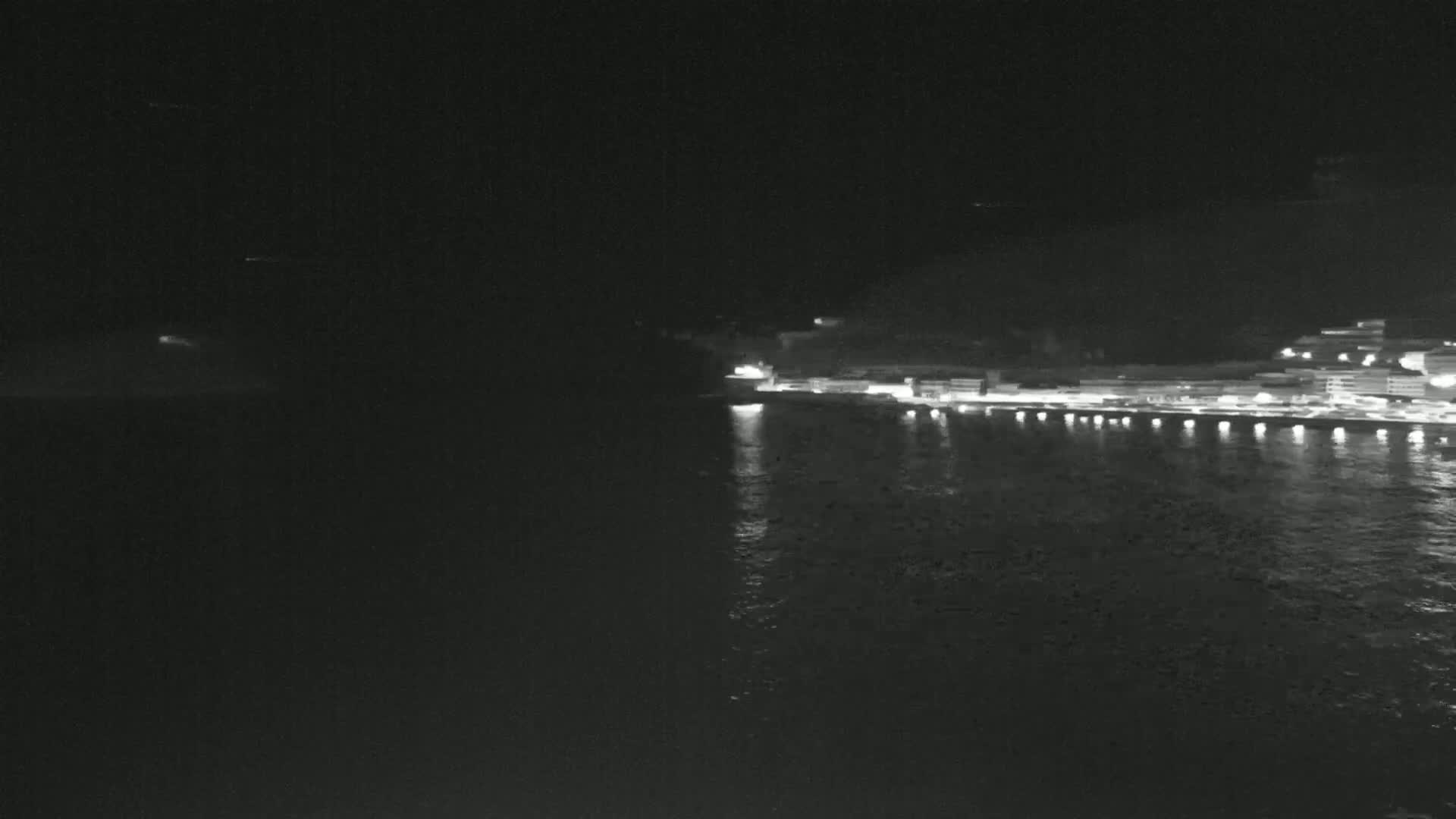 A nighttime, low-angle view shows a dark body of water reflecting the lights of a waterfront town situated against a dark hillside.