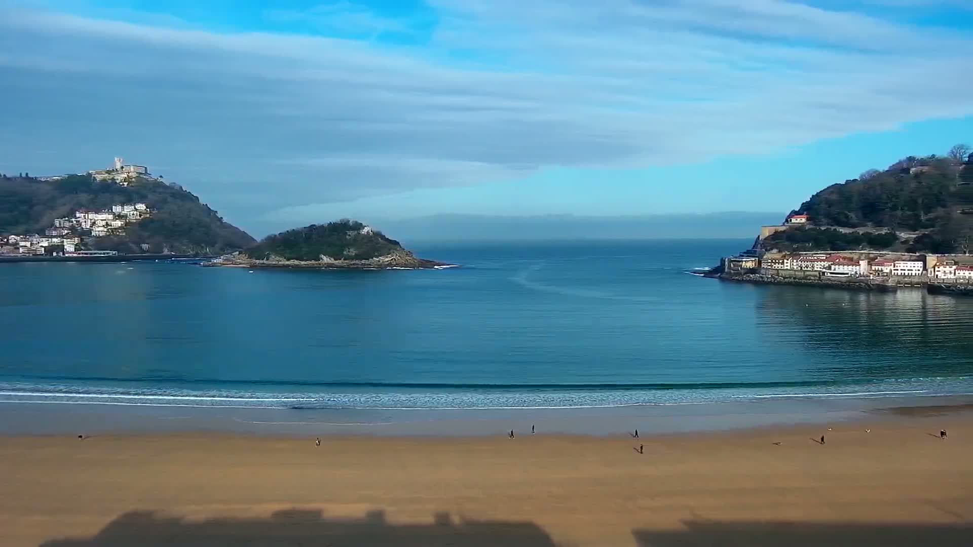A sandy beach with a few people on it faces a calm bay under a mostly clear sky, with hills and towns visible on either side of the bay.