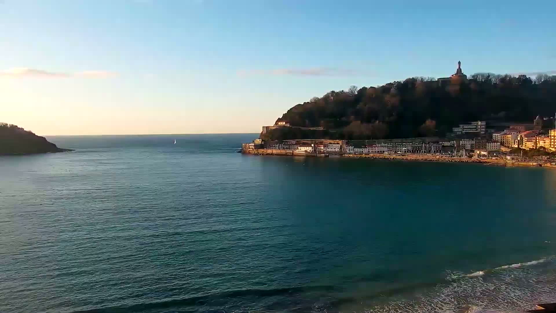 A tranquil bay with a town nestled at the base of a tree-covered hill under a clear, sunny sky.