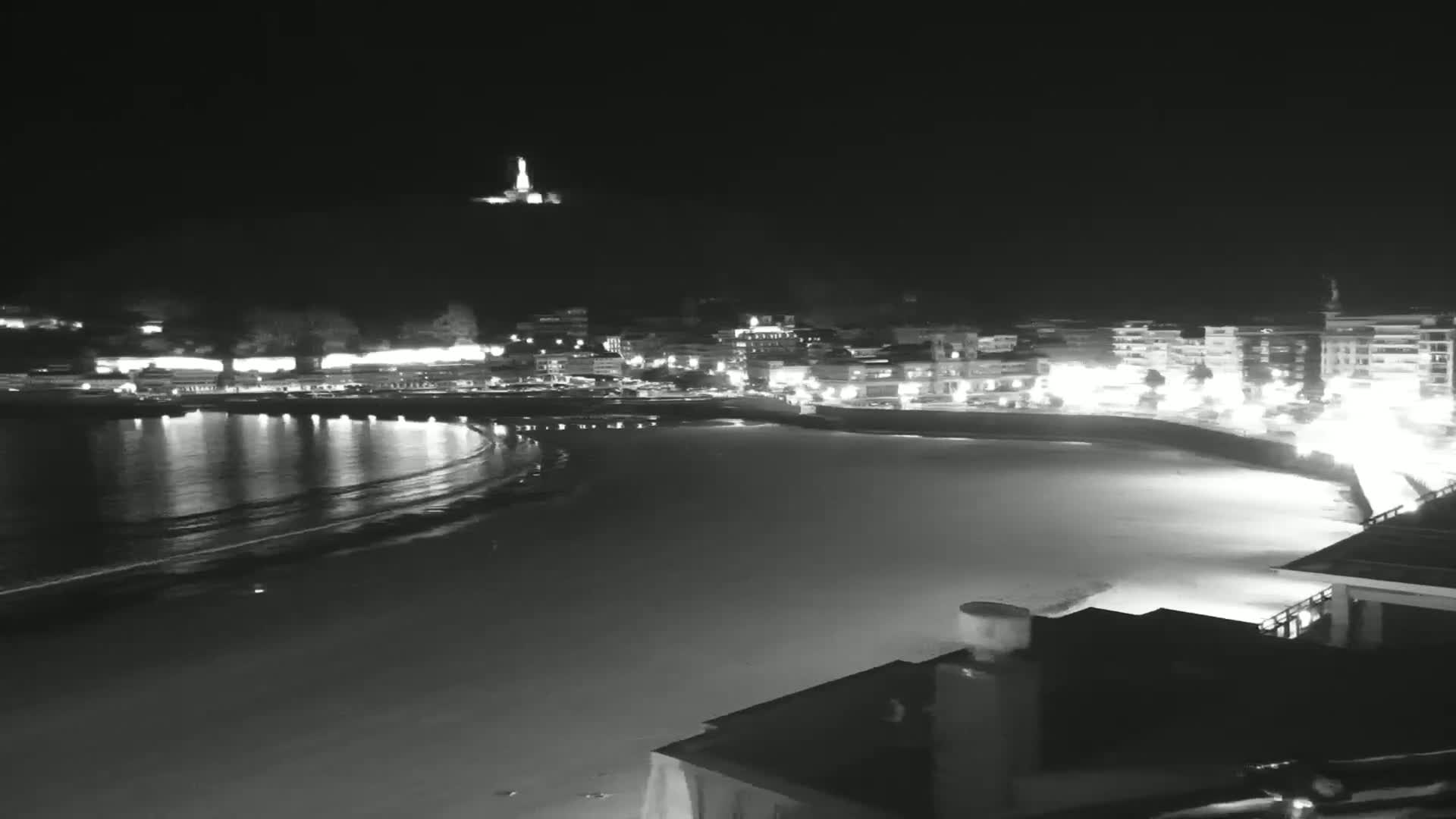 A nighttime, black and white view of a coastal town illuminated with lights, featuring a calm, dark ocean with gentle waves lapping a sandy shore.