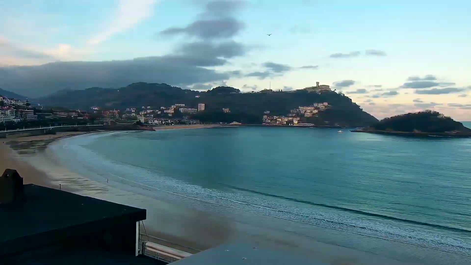 A sandy beach curves around a calm bay, meeting a town nestled against a hillside under a partly cloudy sky.