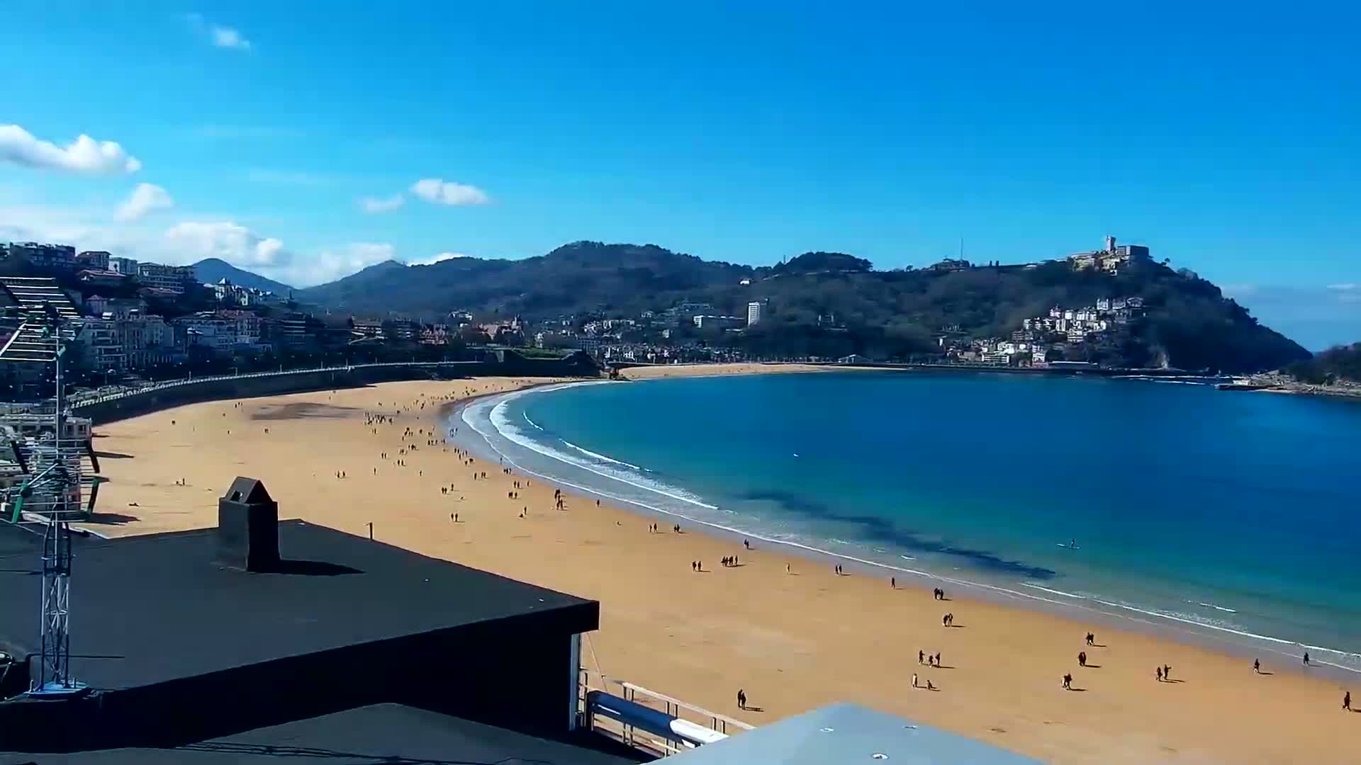 A sandy beach curves around a calm, blue bay under a clear, sunny sky, backed by a town nestled against green hills.