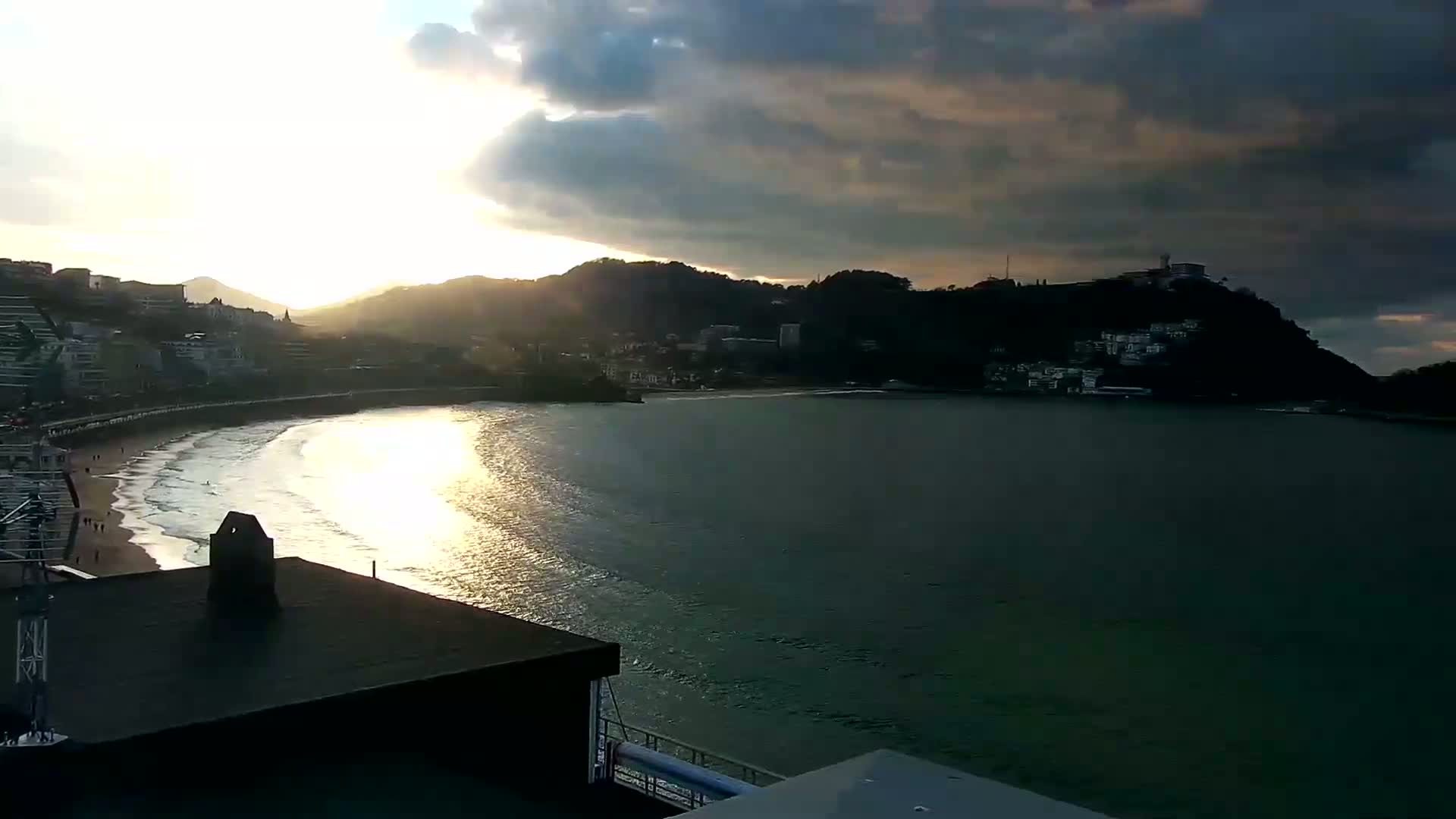 A coastal town sits beneath a partly cloudy sunset sky, with a sandy beach and calm waves lapping the shore.