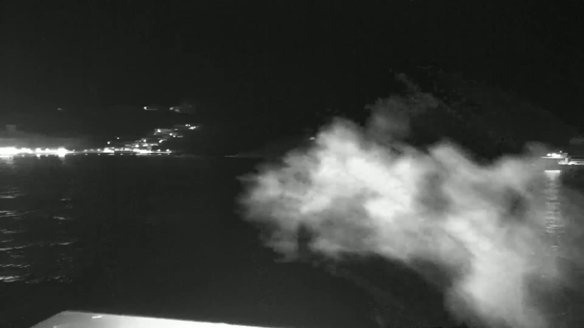 A nighttime, low-light view shows a large plume of white vapor or mist over dark water, with a distant, faintly lit shoreline in the background.