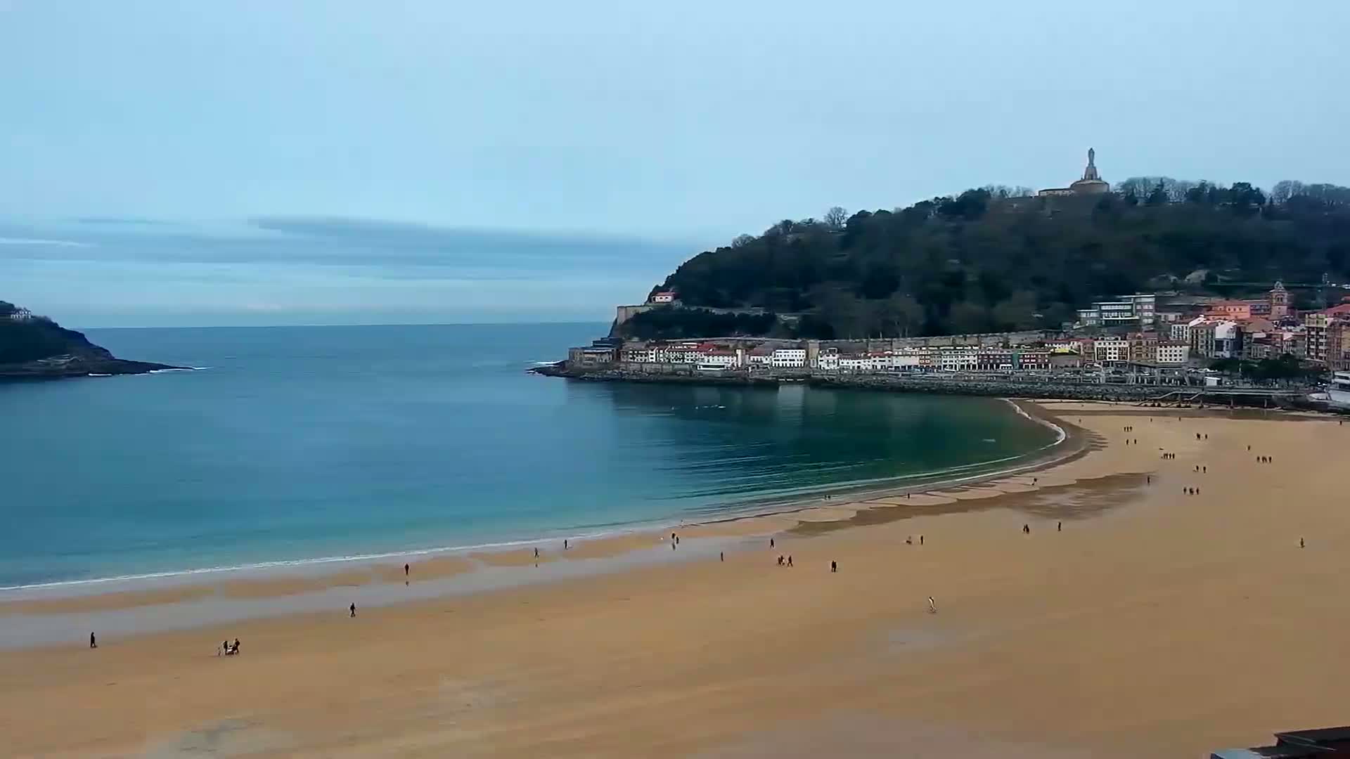 A long sandy beach curves around a calm bay, with a town nestled against a hillside under a cloudy sky.