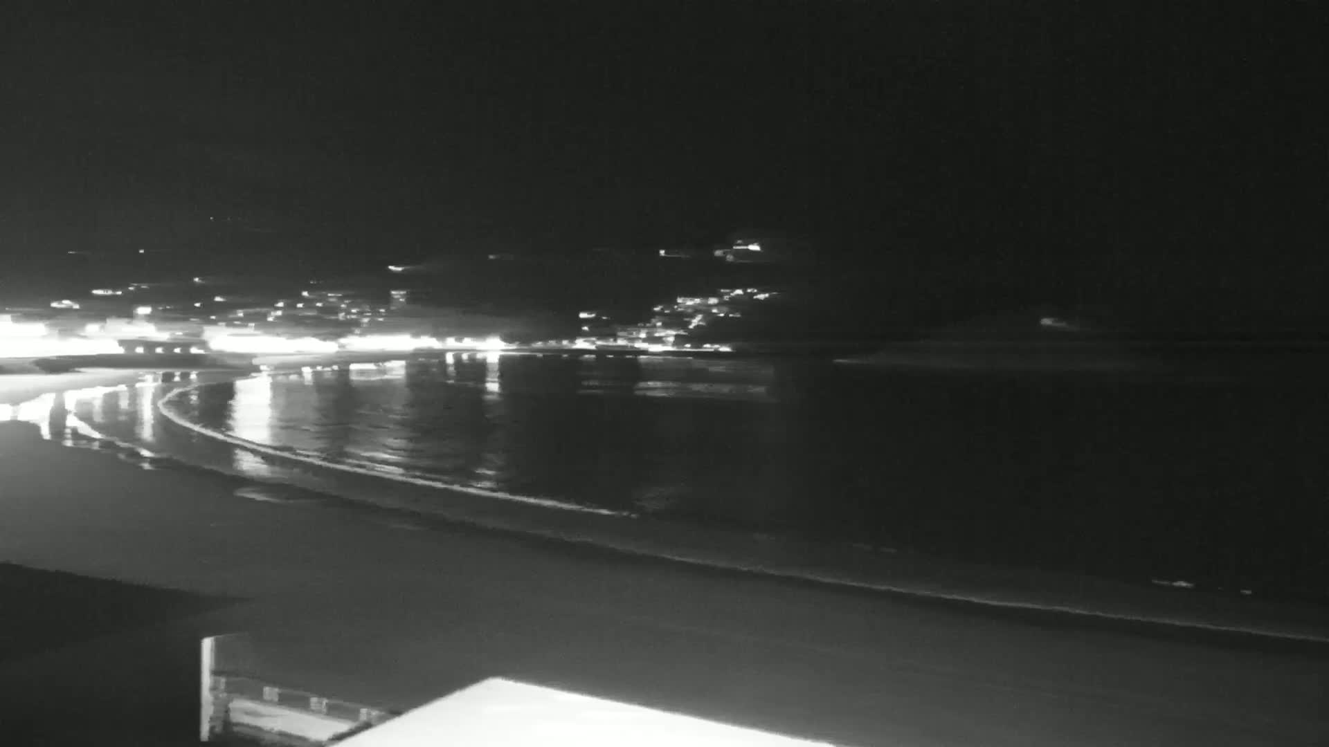 A nighttime view of a dark beach with gentle waves lapping the shore, under a clear, dark sky, and a town's lights visible in the distance.