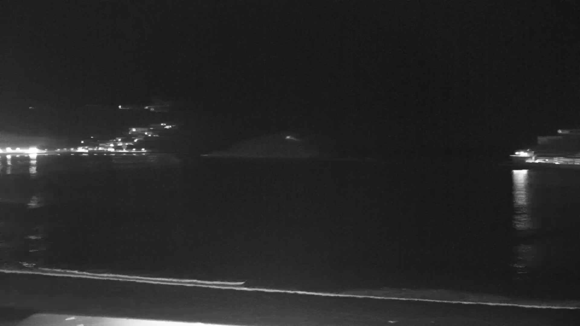 A nighttime view of a calm body of water with faintly lit buildings in the distance and a dark shoreline in the foreground.