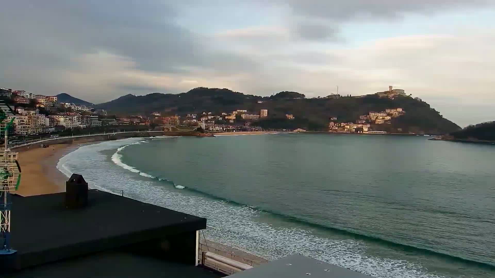 A sandy beach with gentle waves curves around a bay, bordered by buildings nestled against a hillside under a mostly cloudy sky.
