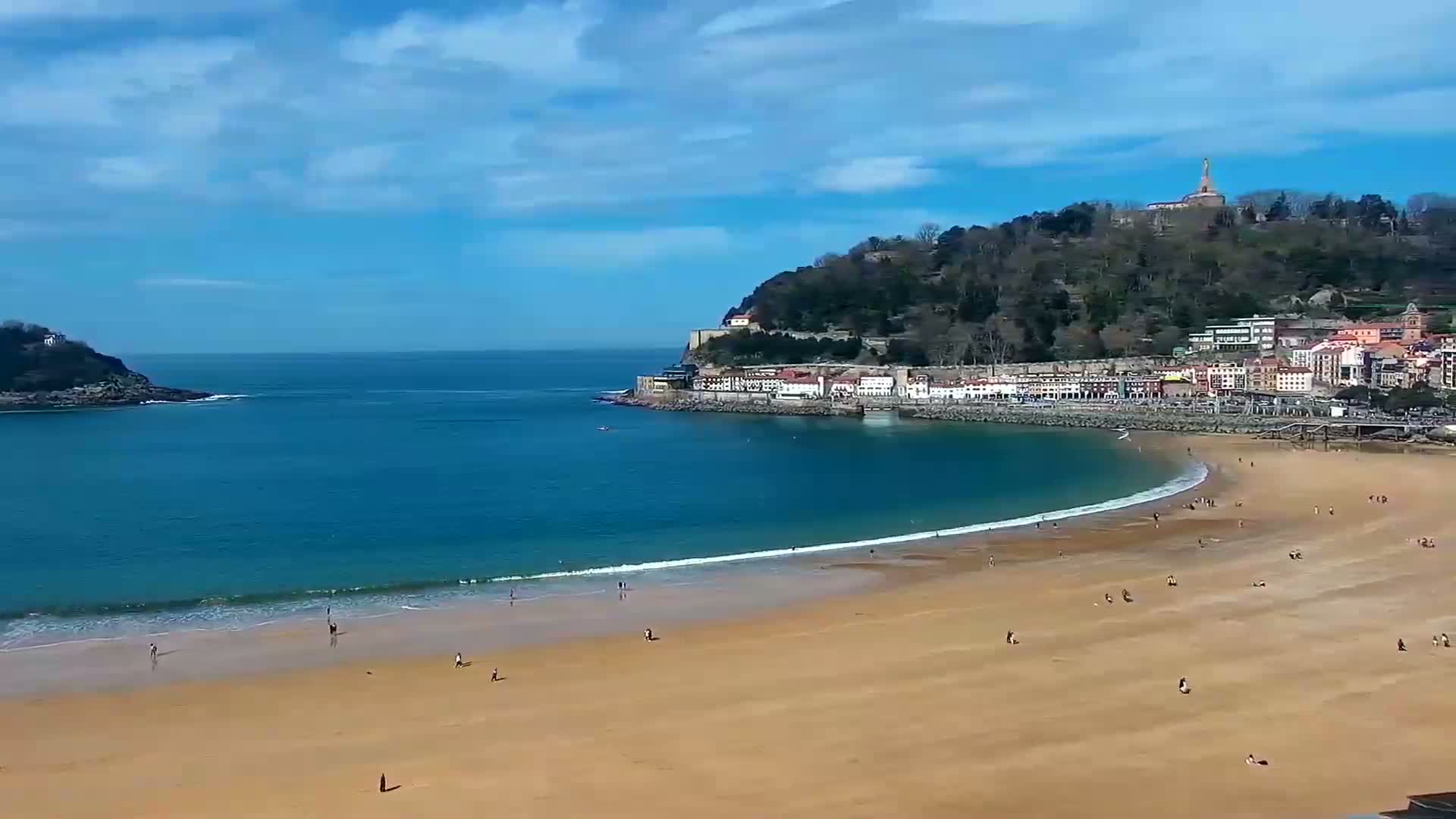 A sandy beach curves around a bay of blue water, bordered by a town nestled on a hillside under a mostly sunny sky.