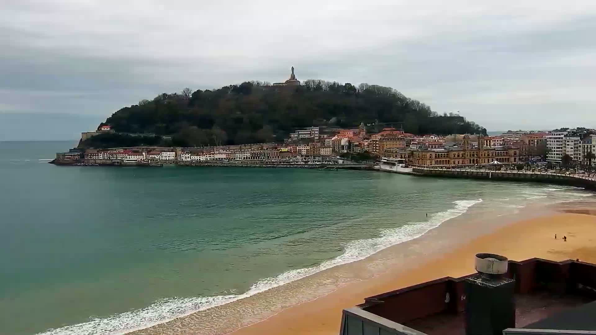 A coastal town nestled against a green hill with a statue atop it, overlooks a sandy beach lapped by gentle waves under an overcast sky.