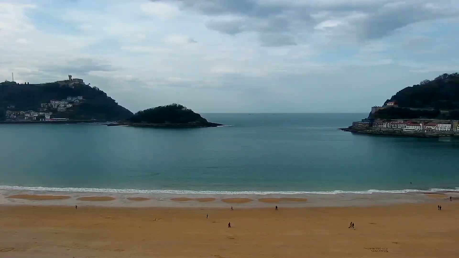 A sandy beach with a few people on it stretches out to a calm bay, which is flanked by hills with buildings on them under a cloudy sky.