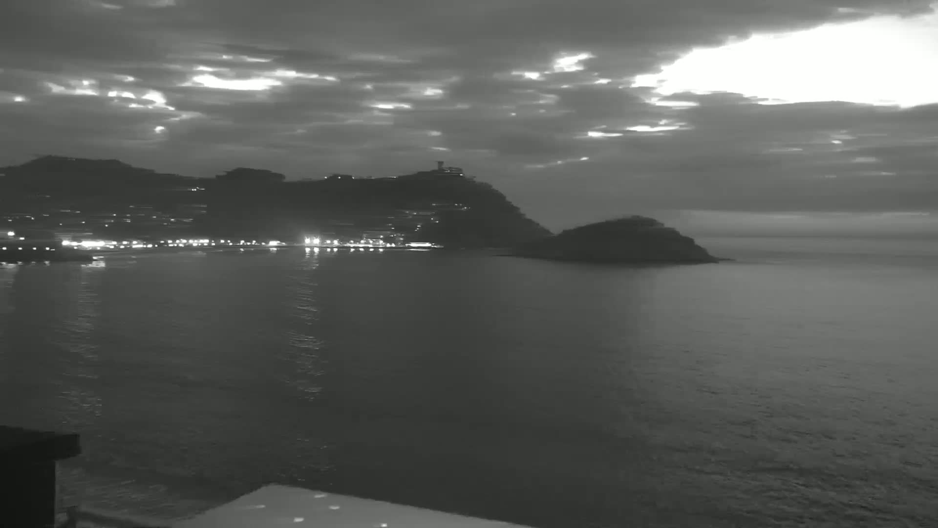 A nighttime coastal scene shows a dark, hilly shoreline with scattered lights, a calm body of water, and an overcast sky with hints of lighter clouds.