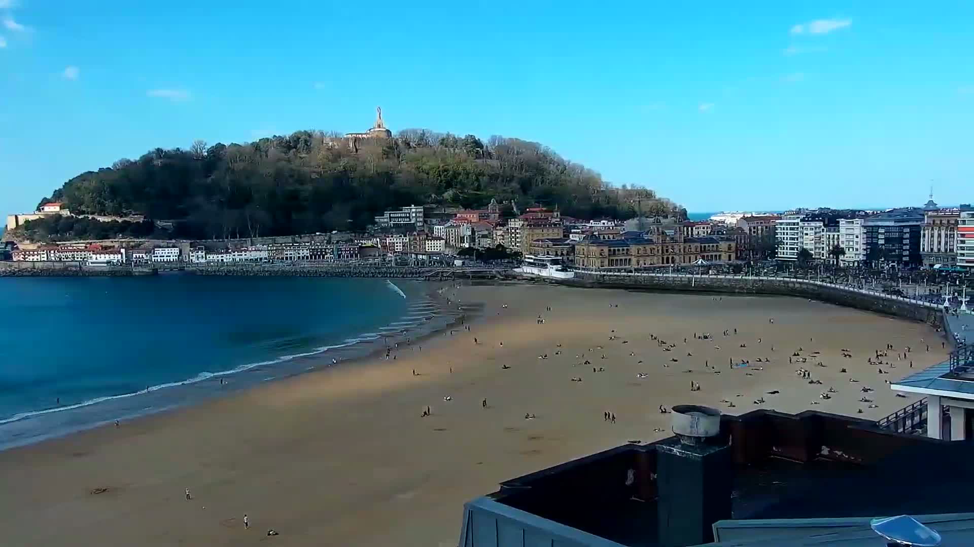 A sandy beach with scattered people stretches beneath a clear blue sky, next to a town nestled against a green hill topped with a monument.