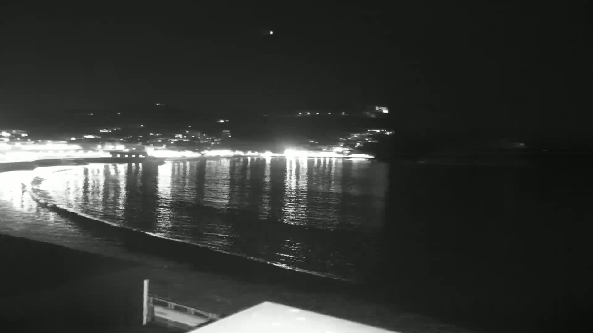 A nighttime view of a calm body of water reflecting the lights of a town across the shore, under a clear sky.