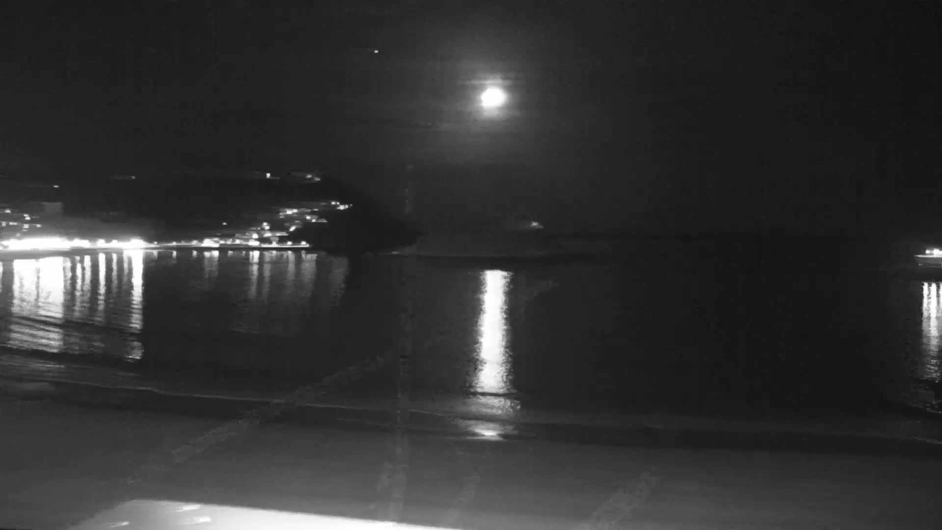 A bright moon shines over a dark body of water reflecting the lights of a distant town on a clear night.