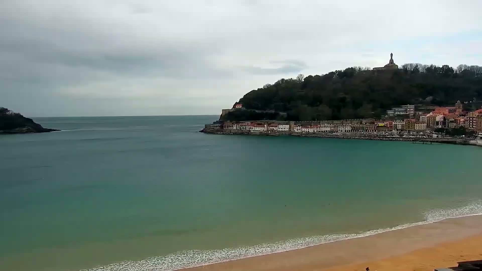 A sandy beach and calm teal ocean meet a coastline with buildings and a hill topped with a statue, under an overcast sky.