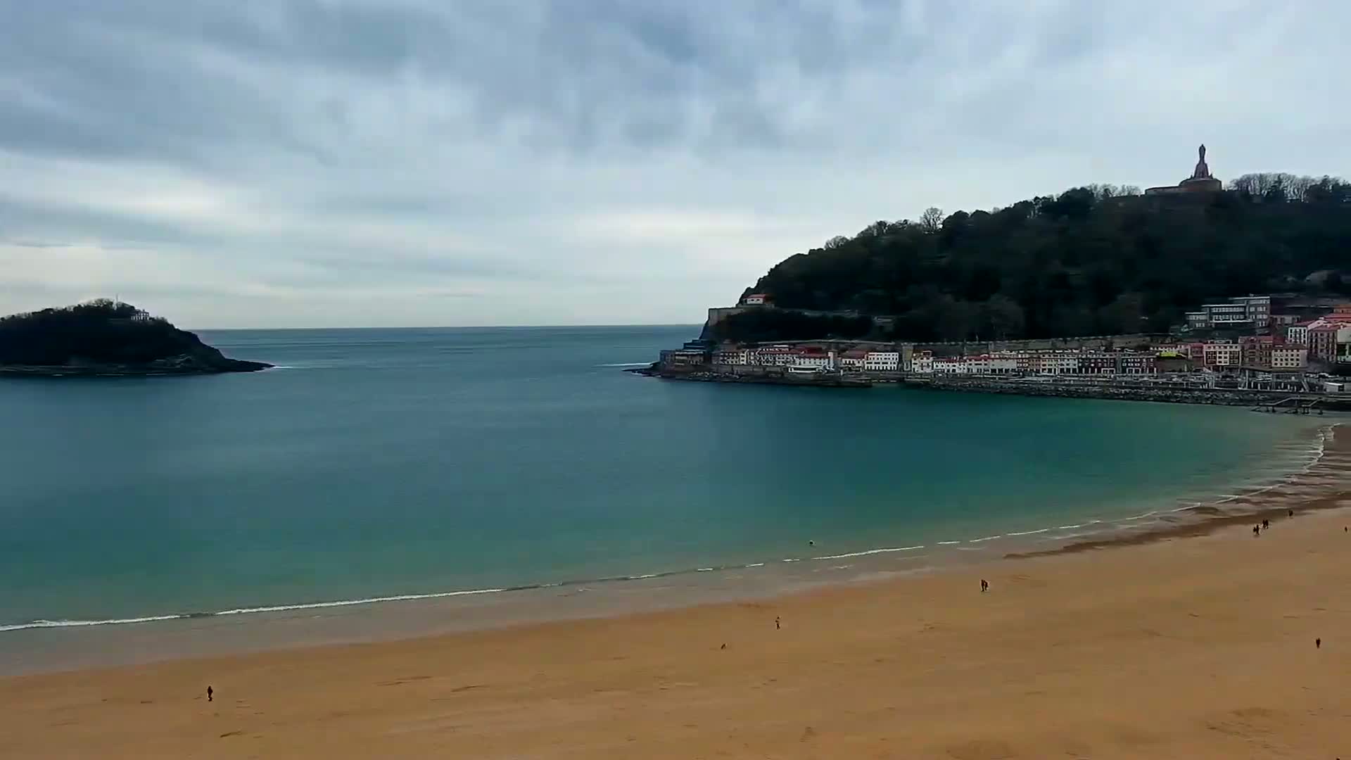 A sandy beach curves around a calm, teal bay, overlooked by a town nestled against a green hill topped with a tall monument, all under a cloudy sky.