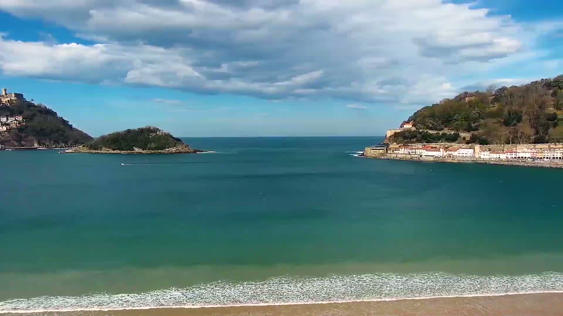 A tranquil bay with a sandy beach and small waves under a partly cloudy sky, flanked by hills with buildings nestled at their bases.